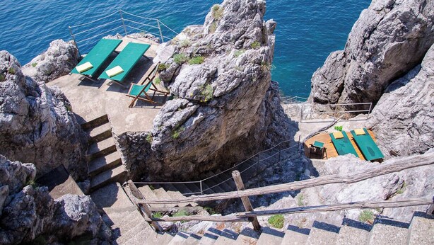 A rocky cliffside with a small outdoor seating area featuring green loungers and a wooden staircase leading down. - Villa Fortino by MyVillaCollection (Maiori)