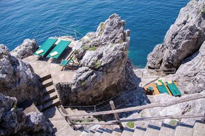 A rocky cliffside with a small outdoor seating area featuring green loungers and a wooden staircase leading down.