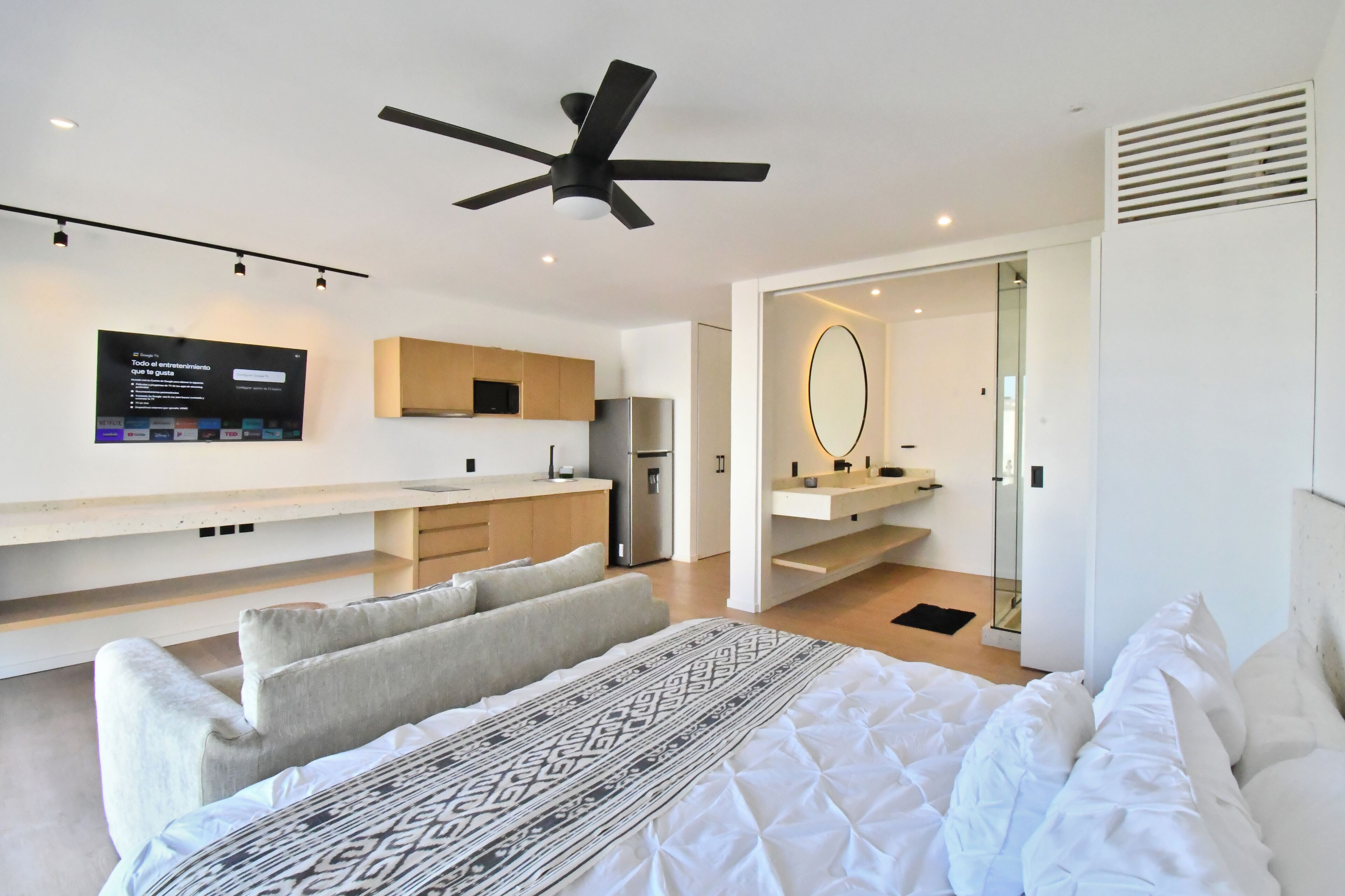 A modern bedroom with a bed, a ceiling fan, a TV, a built-in kitchen area, and a bathroom with a mirror.