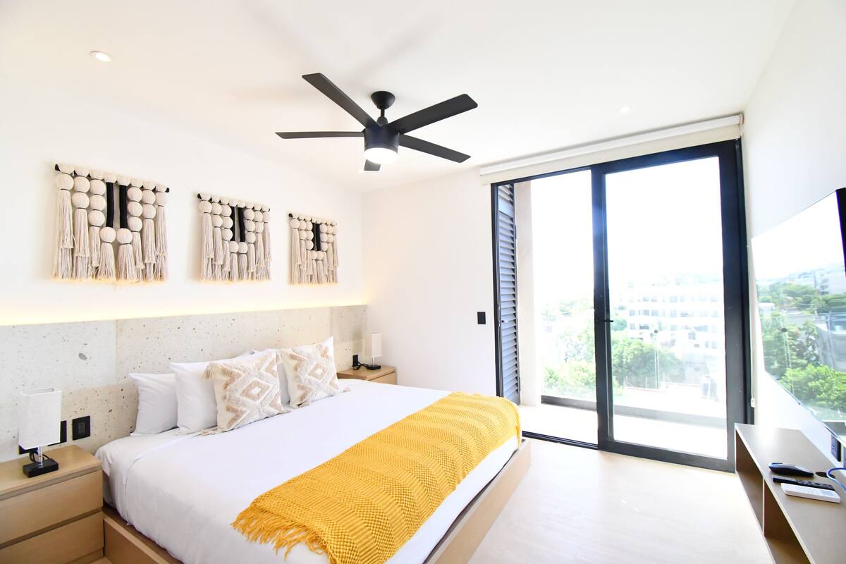 A modern bedroom with a bed, a ceiling fan, a large window, and a view of the city.