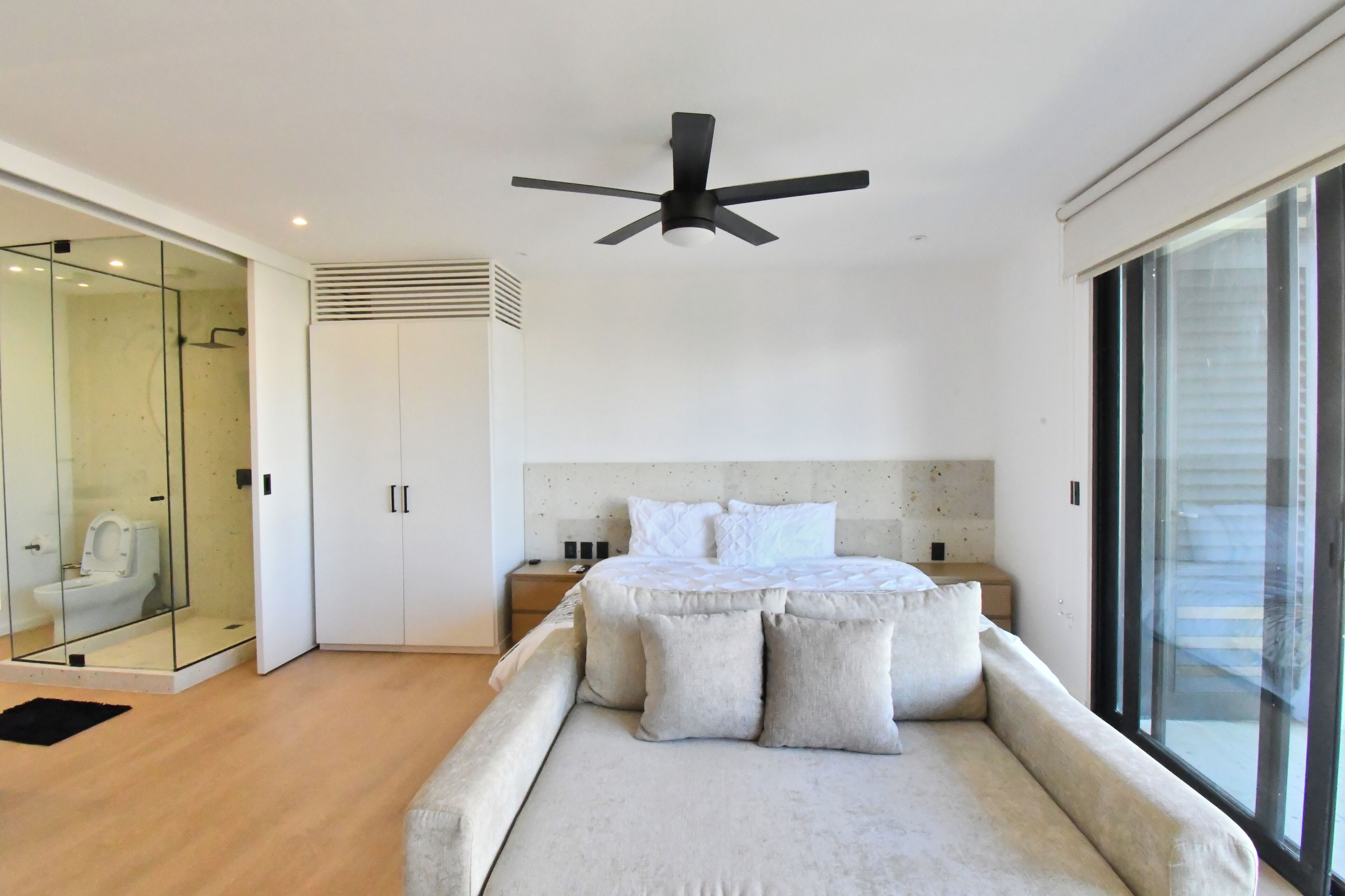 A modern bedroom with a bed, a ceiling fan, a glass shower enclosure, and a large window.