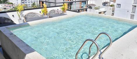 A rooftop infinity pool with clear water, surrounded by modern buildings and a view of the sea.