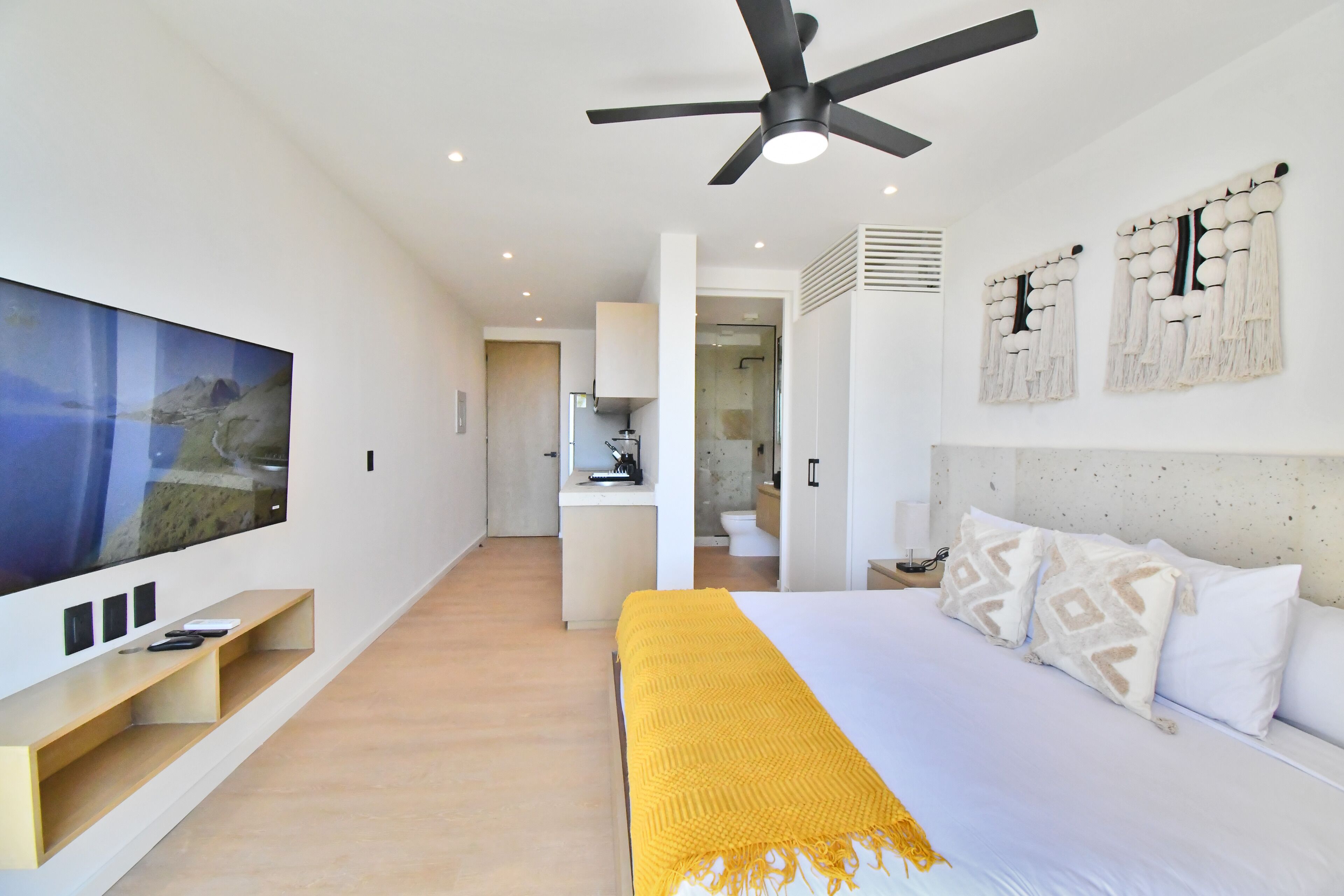 A modern bedroom with a large flat-screen TV, a bed with a yellow throw blanket, a ceiling fan, and a kitchen area visible through an open door.