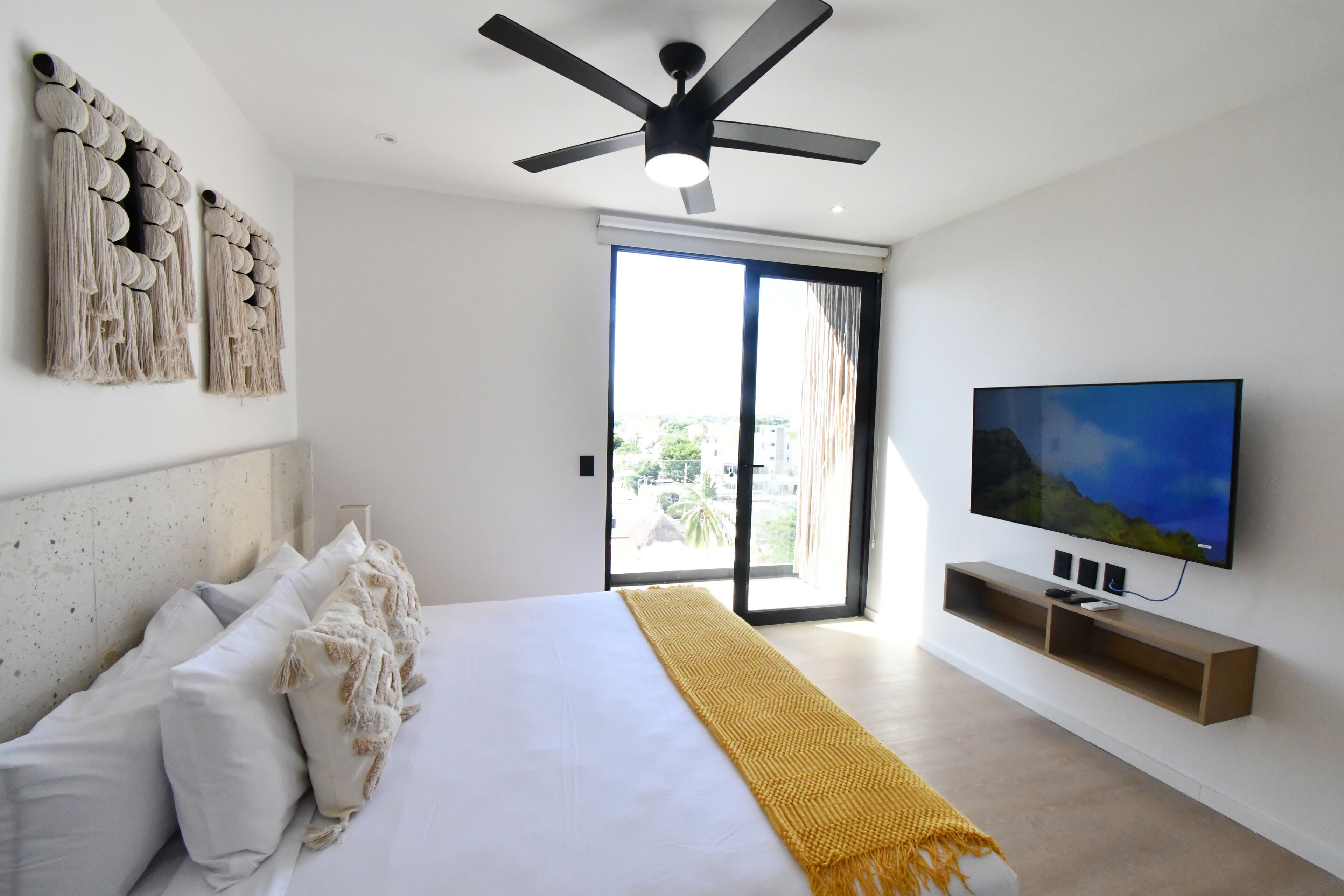 Una habitación de hotel moderna con una cama grande, ventilador de techo, televisor montado en la pared y un balcón con vistas a la ciudad.