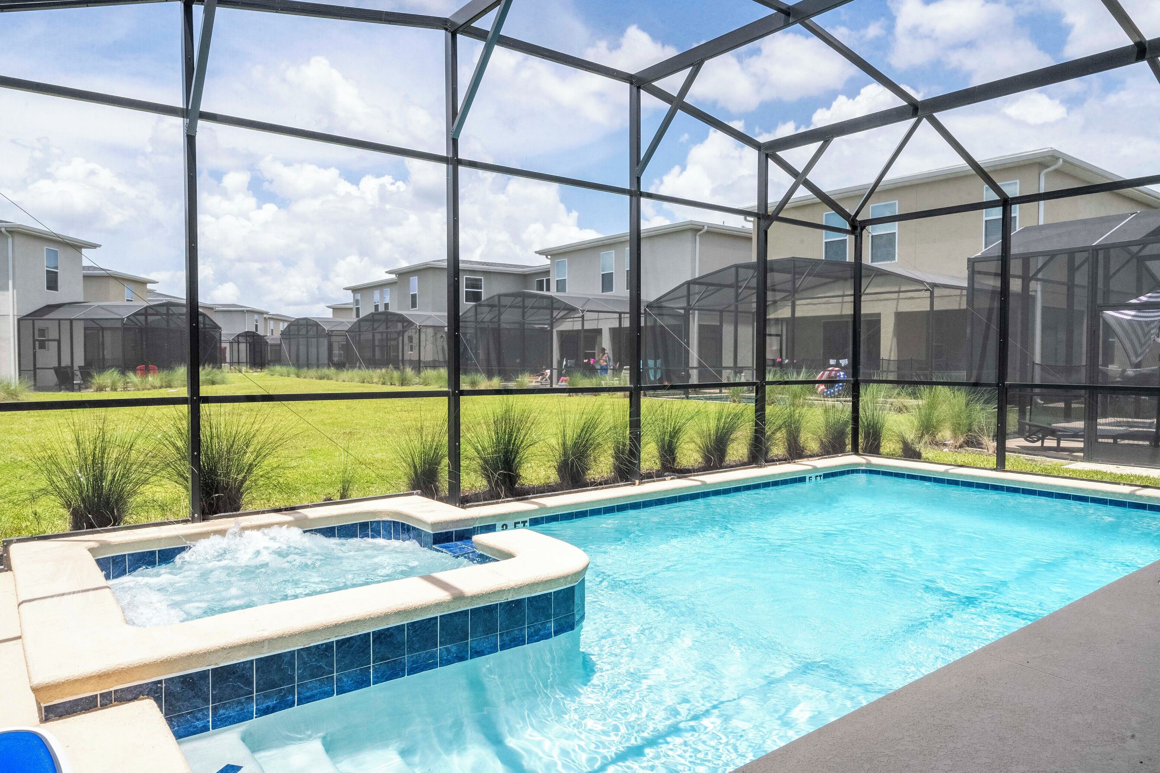 A swimming pool with a hot tub, surrounded by a covered area with a view of houses and a grassy yard.