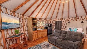 Acogedor interior de una yurta con sofá, televisor, estanterías de madera y zona de cocina.