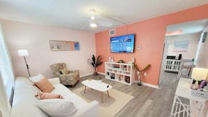 Ein Wohnzimmer mit weißem Sofa, einem kleinen Couchtisch, Deckenventilator, Fernseher an der Wand und einer im Hintergrund erkennbaren Küchenzeile.