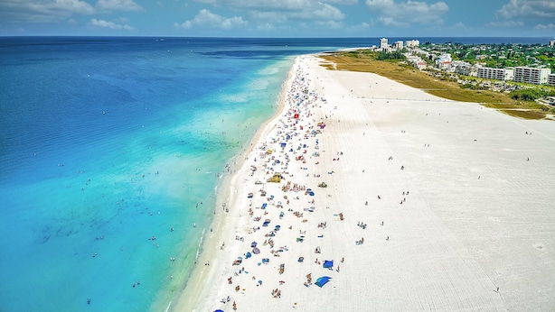 Ein Strand mit einer weiten, hellen Sandfläche, klarem türkisblauem Wasser und einigen Gebäuden im Hintergrund.