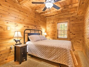 A wooden cabin bedroom with a bed, two bedside lamps, a ceiling fan, and a window.