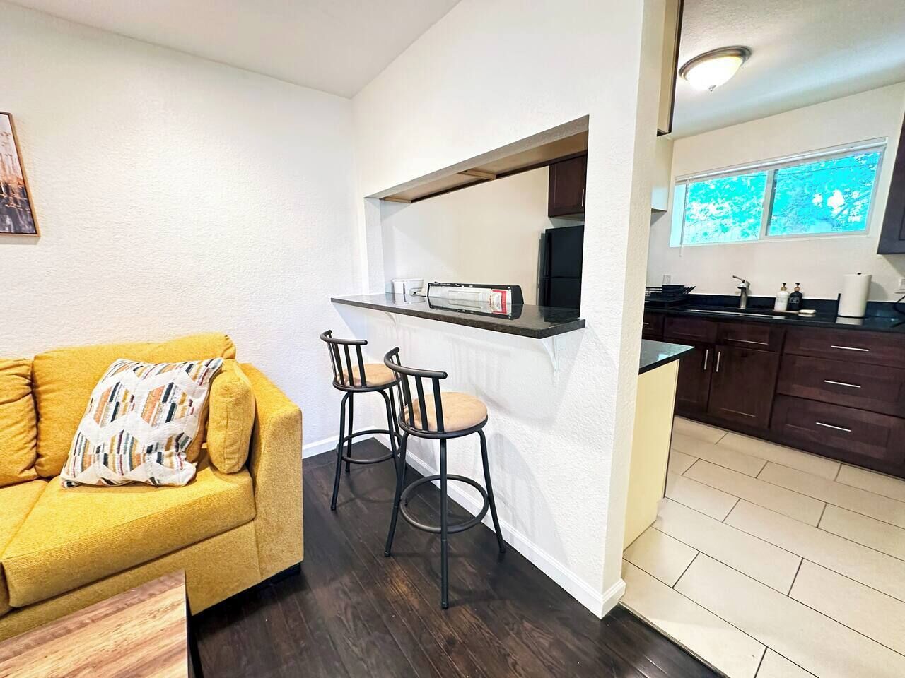 Un espacio habitable pequeño con un sofá amarillo, dos taburetes tipo bar, una zona de cocina con armarios de madera oscura y una ventana con vistas.