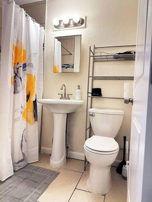 A bathroom with a pedestal sink, a toilet, a shower curtain with a floral pattern, a mirror with lights, and a metal shelf with toiletries.