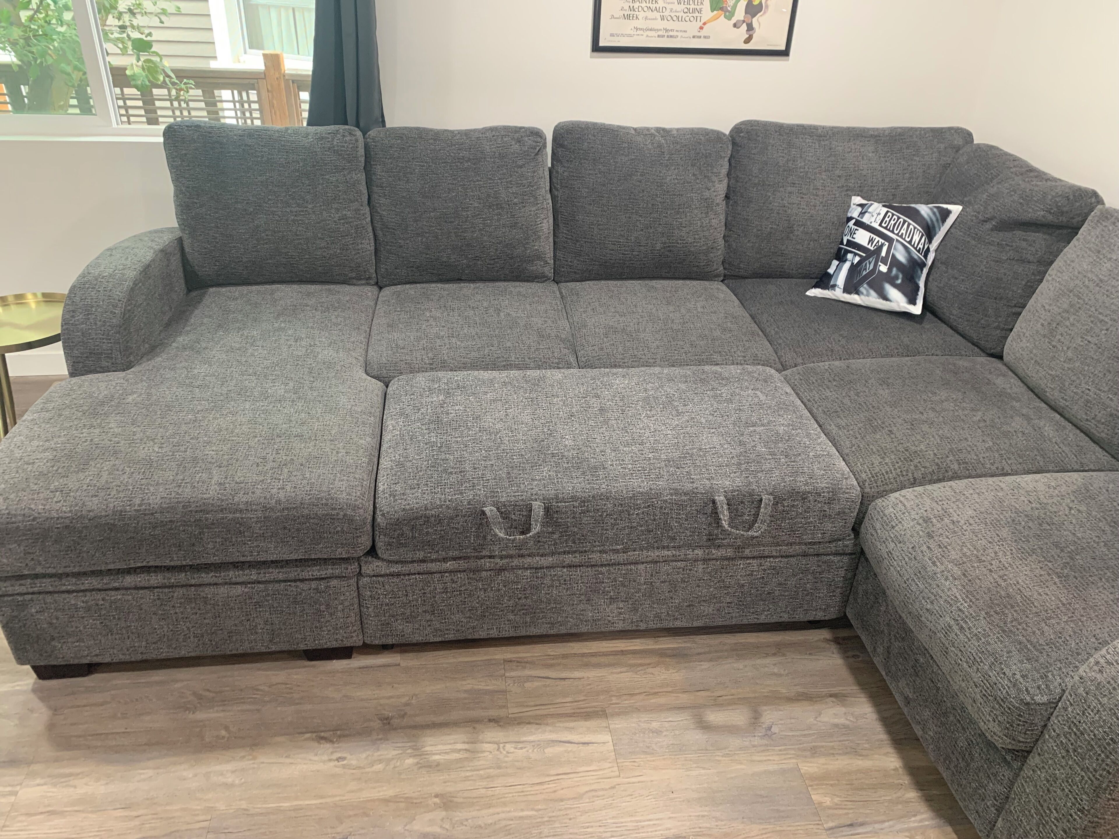 A grey sectional sofa with a small ottoman, a framed poster on the wall, and a window with a view of greenery.