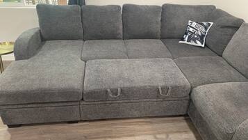 A grey sectional sofa with a small ottoman, a framed poster on the wall, and a window with a view of greenery.