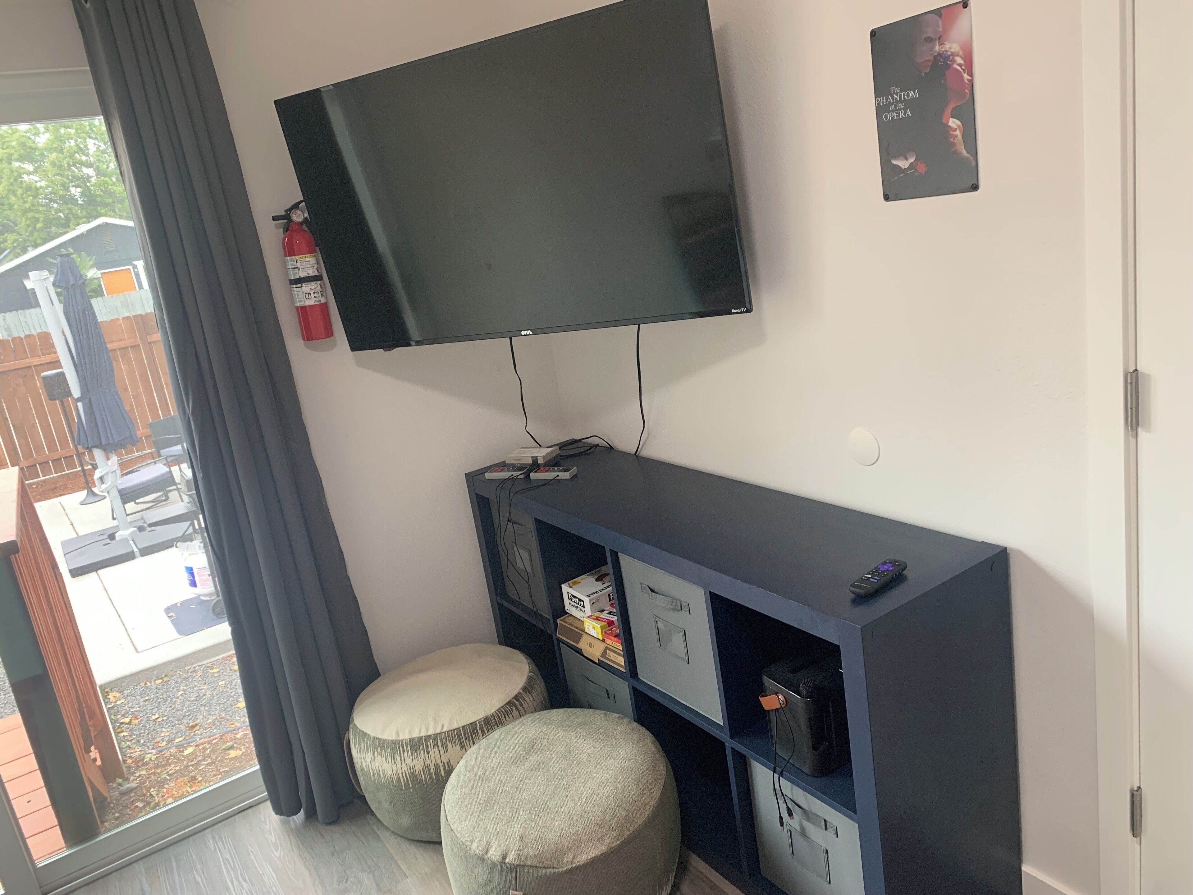 Un saló amb televisió de pantalla plana penjada a la paret, un moble blau d'entreteniment amb espais d'emmagatzematge, dos otomans rodons i un extintor a la paret.
