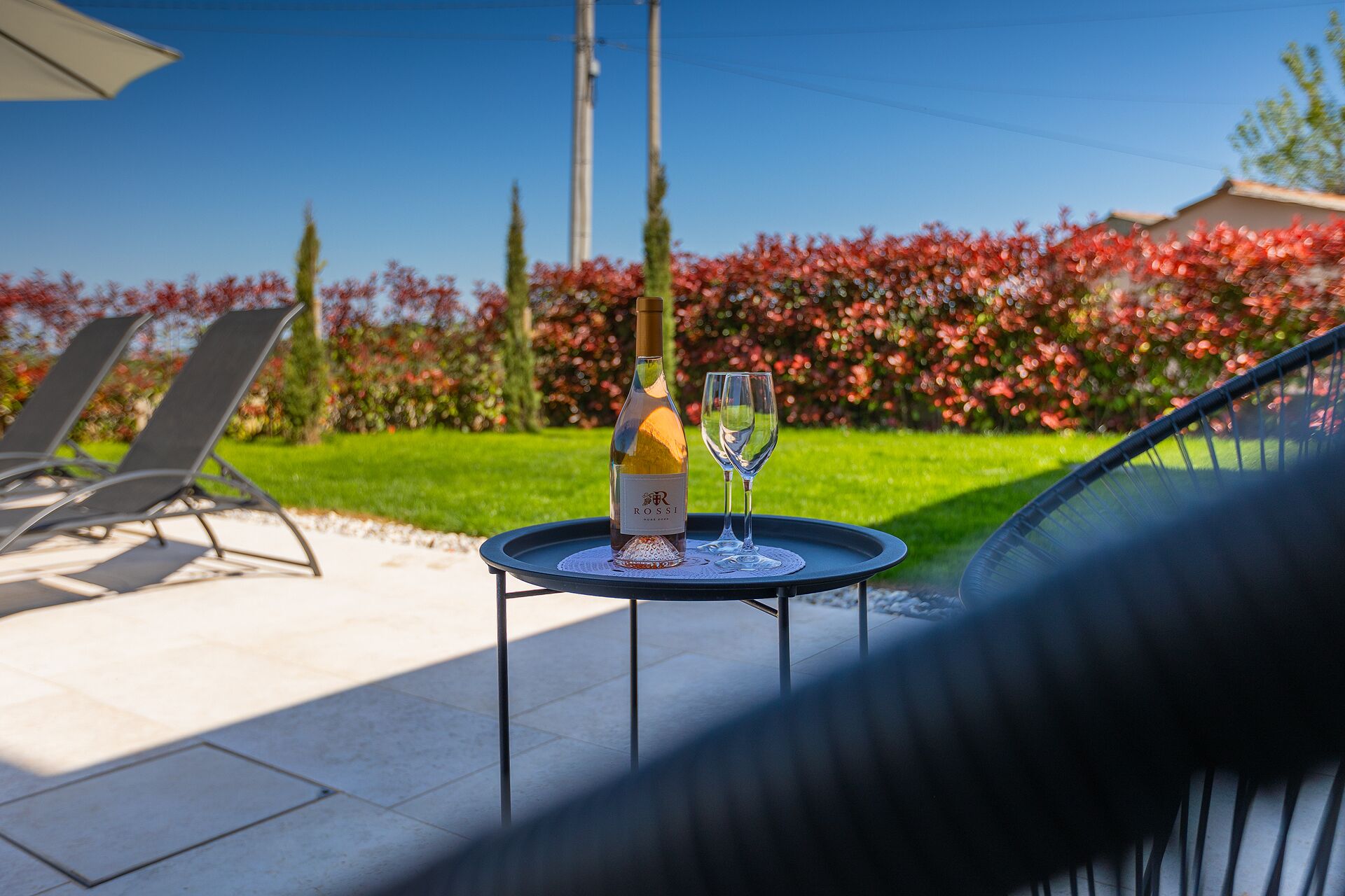 A table with a bottle and a glass of wine, a chair, and a view of a garden with red flowers and a clear sky.