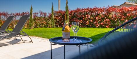 A table with a bottle and a glass of wine, a chair, and a view of a garden with red flowers and a clear sky.