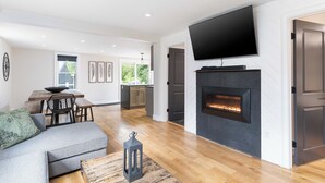 A modern living room with a fireplace, a flat-screen TV, and a dining area with a wooden table and chairs. - ALL NEW, walk to North Conway Village + Railroad, fire pit (Conway)