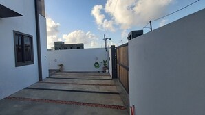 A narrow concrete pathway flanked by white walls, leading to a building with a window and a potted plant.