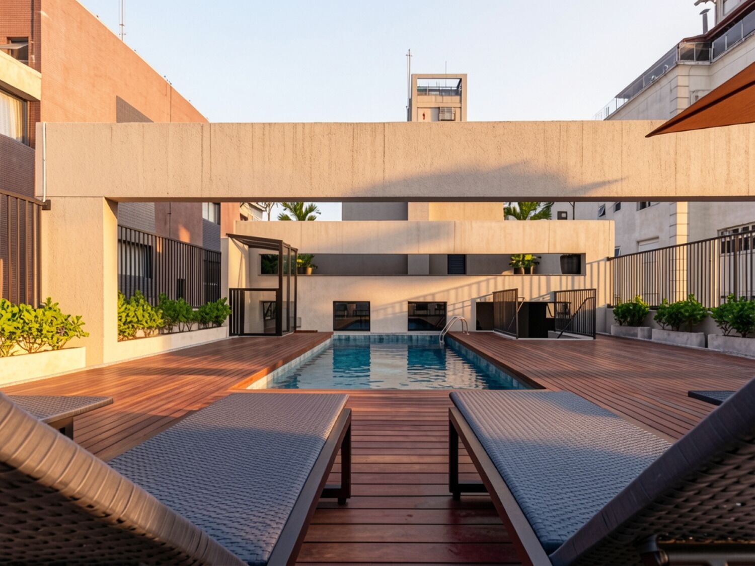 A modern outdoor pool area with wooden decking and lounge chairs, surrounded by buildings with a minimalist architectural style.