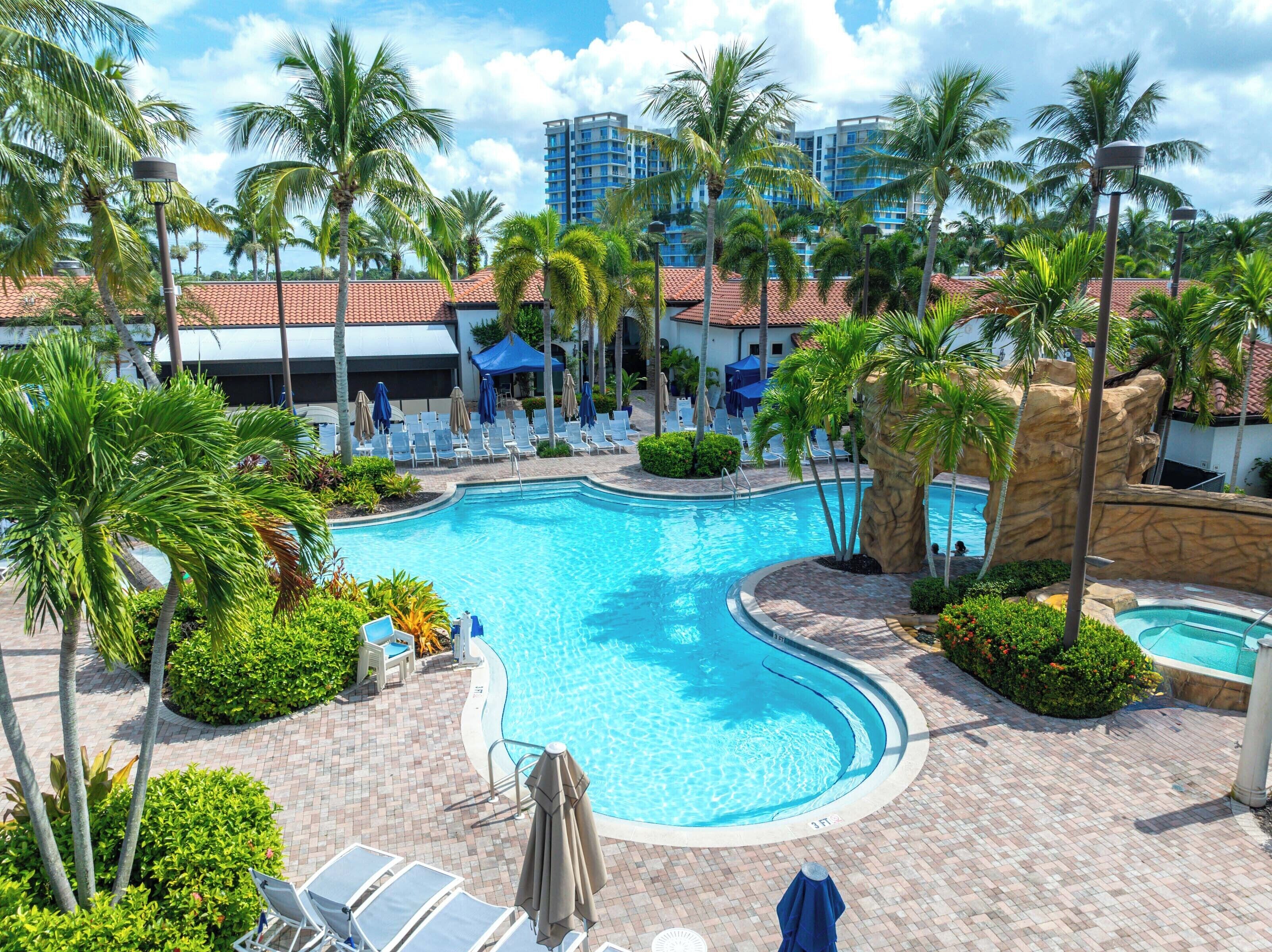 A resort pool area with a large pool, lounge chairs, and palm trees.