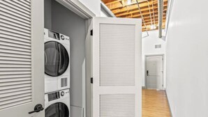 Un lavadero con lavadoras y secadoras apiladas, piso de madera y una puerta blanca que da a otra habitación.