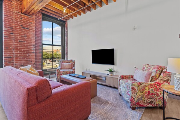 Olohuone, jossa on tiiliseinä, suuri ikkuna, litteänäyttöinen televisio, punainen sohva, kuviollinen nojatuoli ja pieni pöytä, jossa on lamppu.