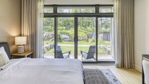 A hotel room with a large bed, a nightstand with a lamp, and a view of outdoor seating and greenery through floor-to-ceiling windows. - 101 Verbier Building 5 (Mont-Tremblant)