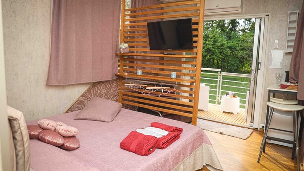 A bedroom with a bed, a television mounted on a wooden slat wall, a balcony with a view of greenery, and a small table with a chair.