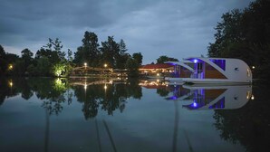 A modern floating structure with a reflective water surface, surrounded by trees and illuminated by ambient lighting.
