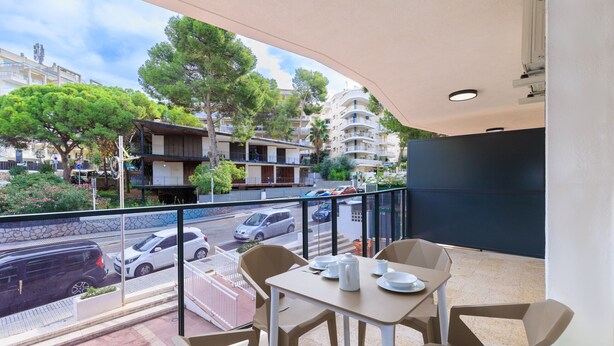 Un balcón con mesa y sillas, con vista a una calle con coches aparcados y edificios.