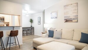 A modern living room with a beige sectional sofa, a kitchen area with wooden flooring, and a dining table.