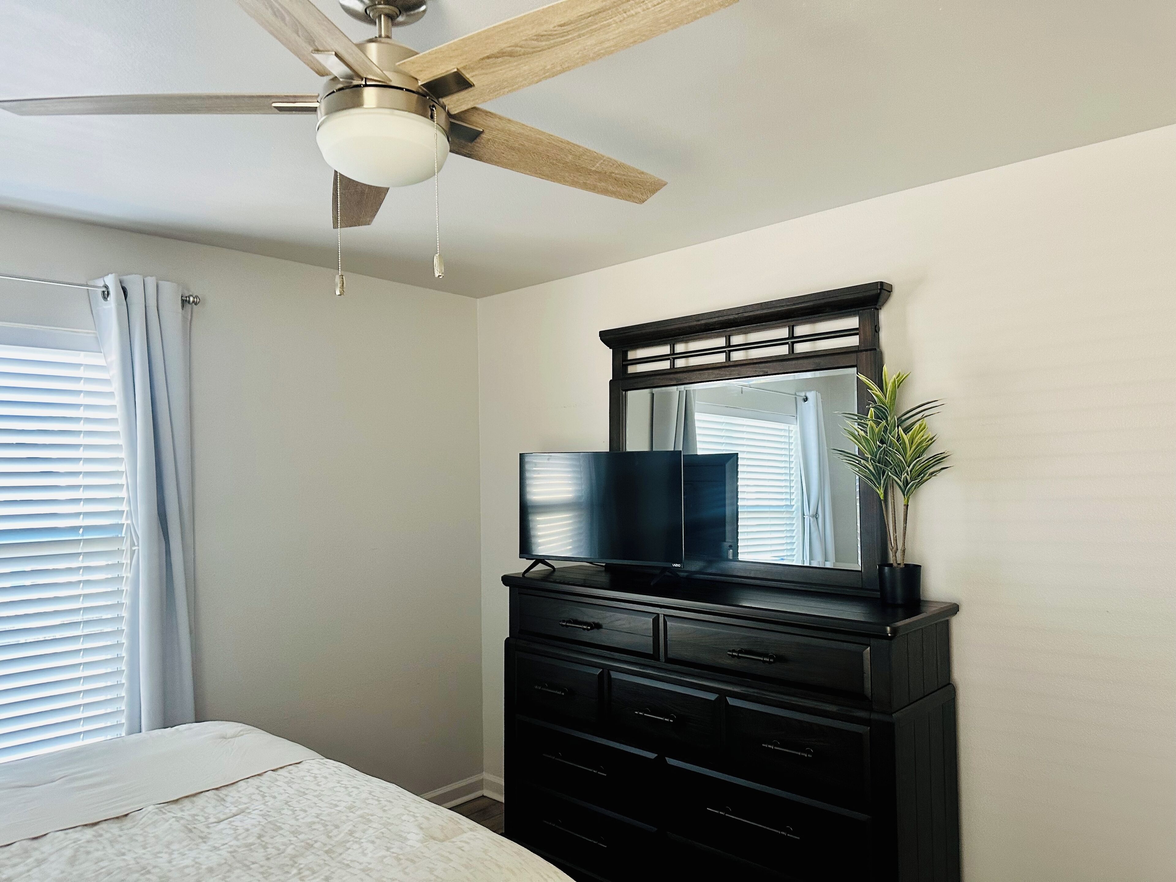 Quarto com uma cama, cômoda preta com gavetas, televisão de tela plana, ventilador de teto e janela com persianas.