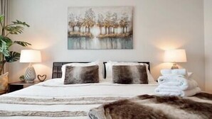 A hotel room with a bed, two bedside lamps, a painting on the wall, and a plant.