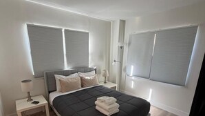 Un dormitorio con cama, mesitas de noche, lámpara y ventanas grandes con persianas.