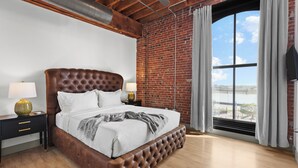 A bedroom with a large bed, wooden headboard, brick walls, and a large window with curtains. - DWTN Riverfront Loft W/D Gated Parking (St. Louis)