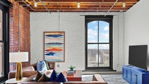 A modern living room with a brick wall, a large window, a sofa, a coffee table, and a TV stand. - DWTN Riverfront Loft W/D Gated Parking (St. Louis)