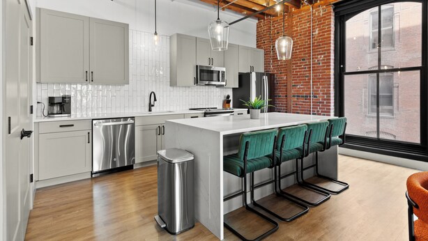 A modern kitchen with white cabinets, stainless steel appliances, and a large island with bar stools. - DWTN Riverfront Loft W/D Gated Parking (St. Louis)