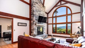 A spacious living room with a stone fireplace, a large window offering a scenic view, and a leather sofa. - Modern Smoky Mountain Cabin for Big Views, Lake Days, and Hot Tub Nights (Bryson City)