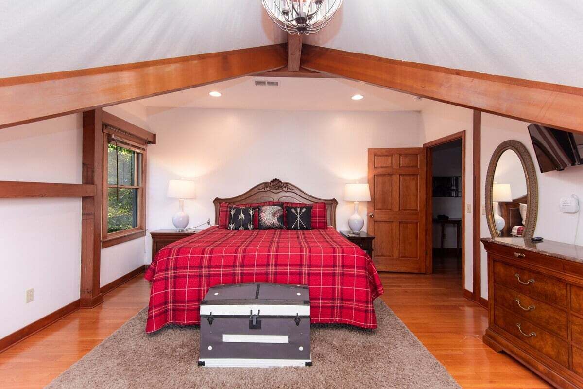 A bedroom with a red plaid bedspread, wooden beams, a wooden dresser, a mirror, and a suitcase on the floor.