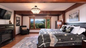 A bedroom with a large bed, a chandelier, a TV, and a view of the outdoors. - Modern Smoky Mountain Cabin for Big Views, Lake Days, and Hot Tub Nights (Bryson City)