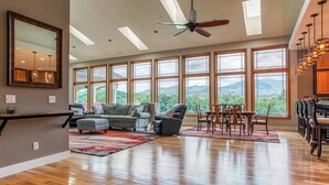 Un salón amplio con un ventanal grande, un ventilador de techo y una zona de comedor con sillas de madera.