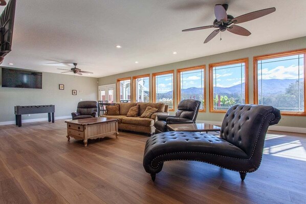Relax in this spacious living room with a high ceiling and large windows offering a view of the surrounding landscape. Enjoy a comfortable seating area with a sofa and armchair, plus a coffee table and flat-screen TV.