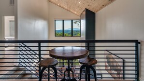 A modern interior with a wooden ceiling, large windows offering a view of the outdoors, and a round table with stools. - NEW Heated Pool & Theater Luxury Getaway|Mtn Views (Sevierville)