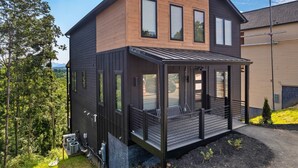 A modern two-story house with a wooden exterior, large windows, and a covered porch with seating. - NEW Heated Pool & Theater Luxury Getaway|Mtn Views (Sevierville)