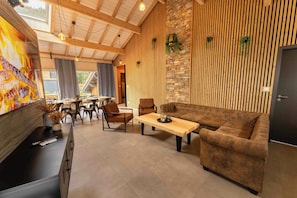 A modern living room with a sofa, coffee table, and chairs. There is a dining area with a table and chairs. The room has wooden beams on the ceiling and a stone wall.