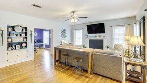 A spacious living area with a wooden floor, a sofa, a coffee table, and a TV mounted on the wall. There is a ceiling fan, a bookshelf, and a small table with stools.