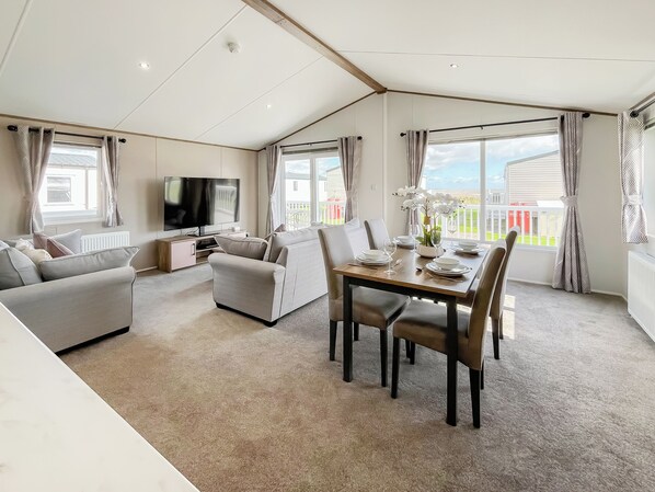A spacious living area with a sofa, a dining table set for four, a television, and a balcony with a view.