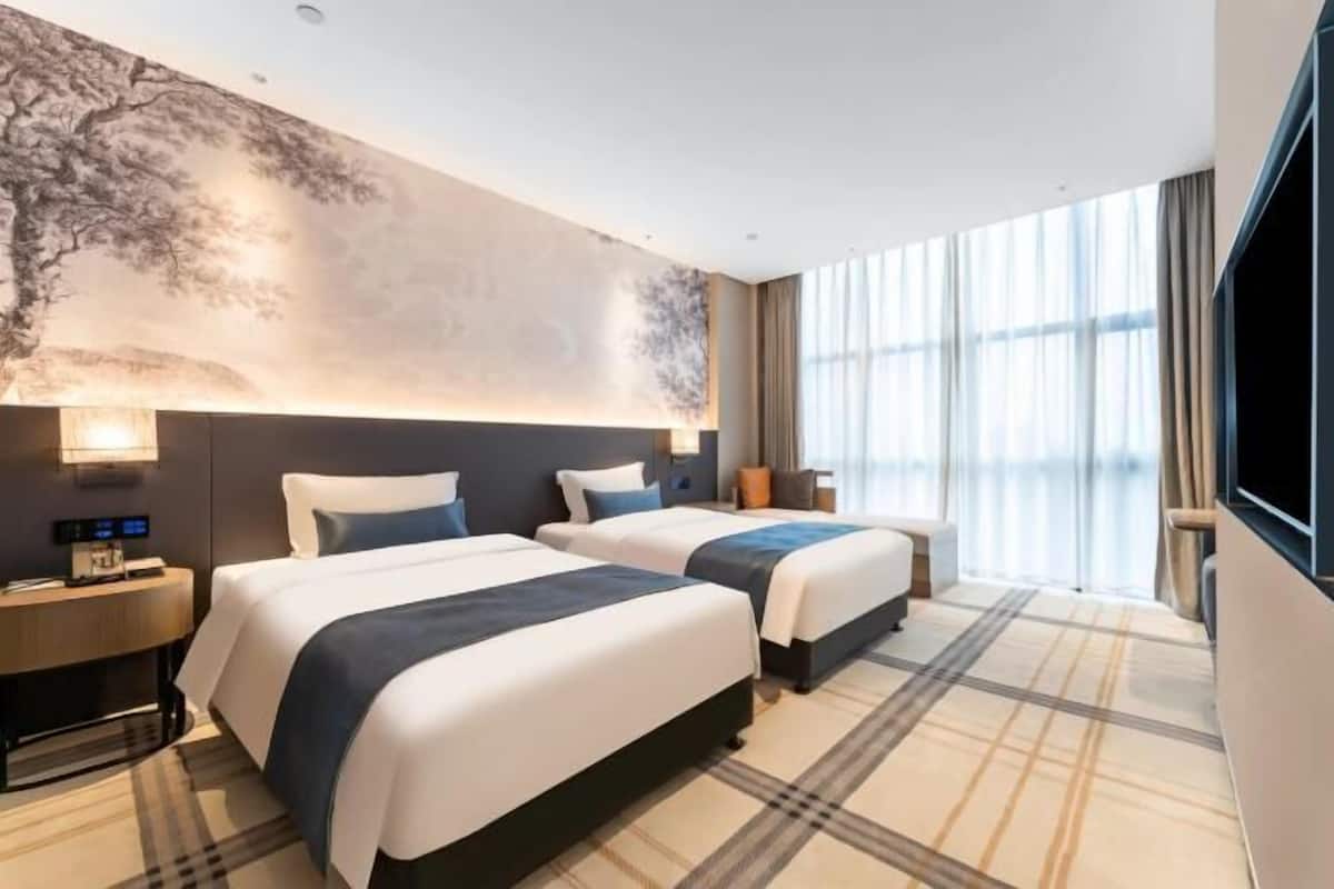 Hotelkamer met twee bedden, een groot raam met gordijnen, een wandtelevisie en een nachtkastje met een lamp.
