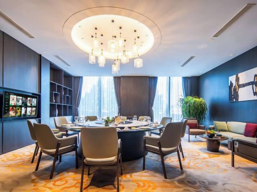 Sala de conferências moderna com mesa redonda, cadeiras, candelabro, tela de televisão e um sofá.