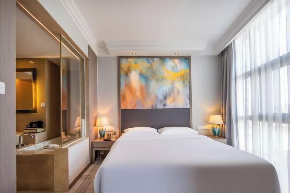 Een hotelkamer met een groot bed, een schilderij aan de muur, een badkamer met een spiegel en een raam met gordijnen.